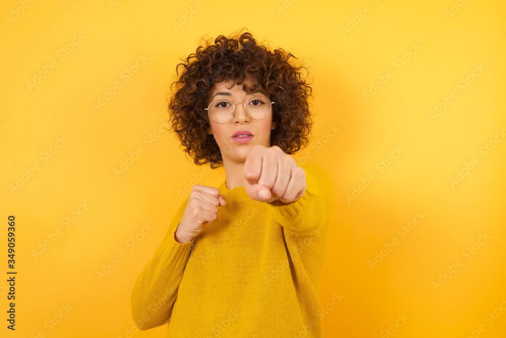Young arab woman with curly hair wearing yellow sweater standing over isolated yellow background Punching fist to fight, aggressive and angry attack, threat and violence Young arab woman with curly hair wearing yellow sweater standing over isolated yellow background Punching fist to fight, aggressive and angry attack, threat and violence
