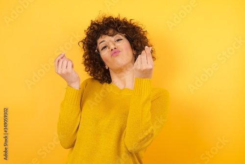Young arab businesswoman with curly hair wearing sweater standing over isolated yellow background doing money gesture with hands, asking for salary payment, millionaire business © Roquillo