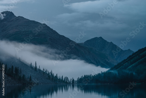 Low cloud above alpine lake. Silhouettes of trees reflected on mountain lake. Firs and pines above calm water in dense fog. Highland tranquil landscape at early morning. Ghostly atmospheric scenery.