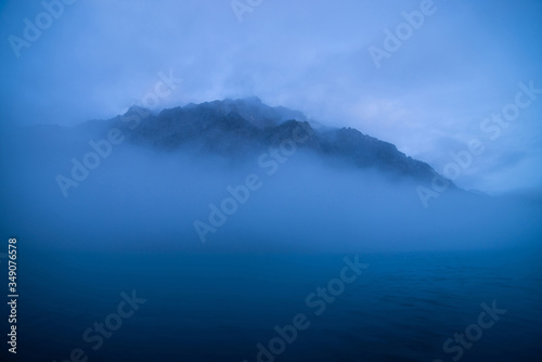 Tranquil minimal landscape with big rocky mountain in middle of water among low clouds in twilight. Wavy sea of blue classic color. Atmospheric scenery with deep blue calm lake and rock in dense fog. © Daniil