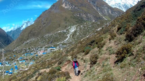 Young man wearing hat and carrying trekking pole hikes uphill in the Langtang valley of Nepal.