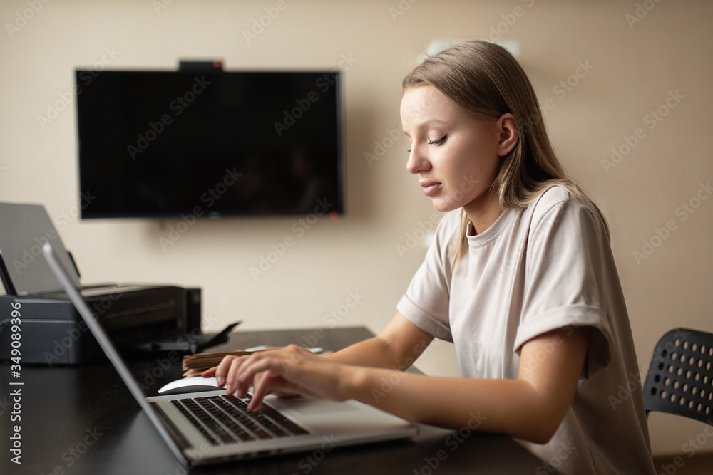 Cute pensive girl is doing work from home typing text on a computer with a notebook and a phone on the table during quarantine and pandemic.