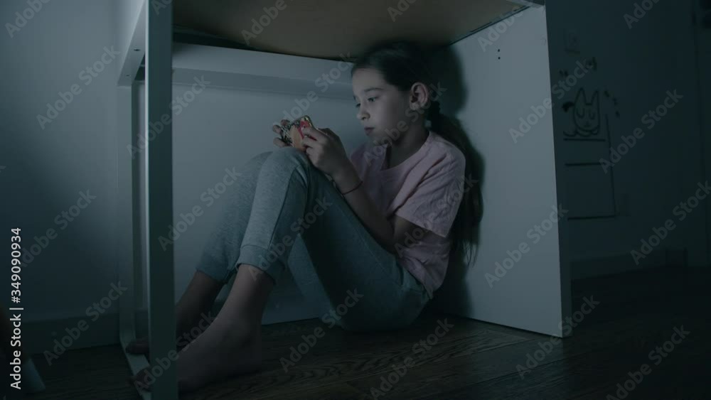 Little girl emotionally plays in smartphone under table at night Little girl emotionally plays in smartphone under table at night