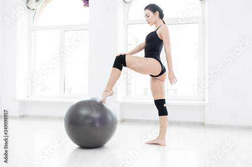 Sexy attractive woman in black sportswear is posing in the studio touching a fitness ball with her toe