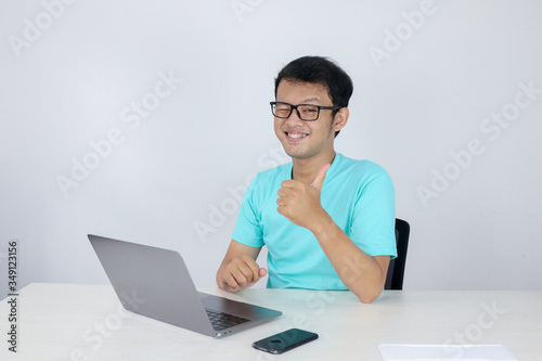 Young handsome asian man showing thumb up while working with laptop. Indonesian man wearing blue shirt. © Reezky