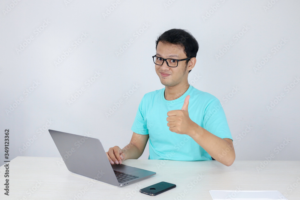 Young handsome asian man showing thumb up while working with laptop. Indonesian man wearing blue shirt.
