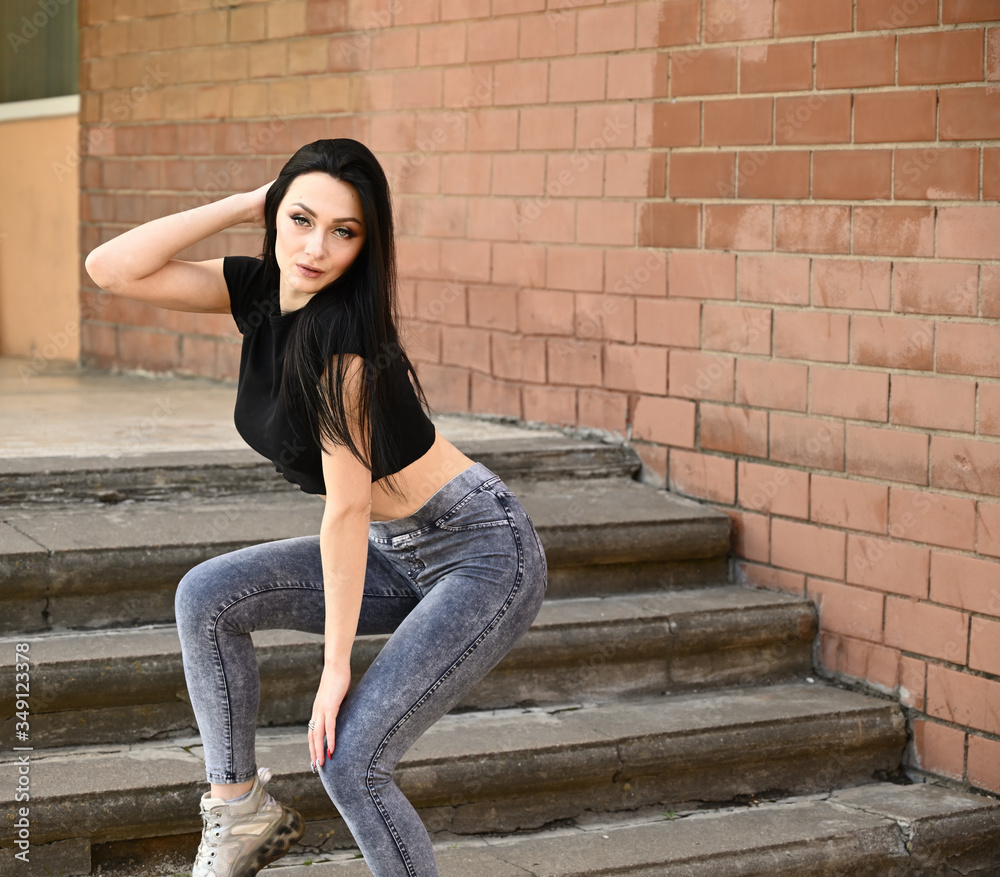 Model posing on the background of the stairs of the building. Full-length portrait in spring weather outdoors, caucasian brunette girl with a slim figure in jeans and a black T-shirt. Model posing on the background of the stairs of the building. Full-length portrait in spring weather outdoors, caucasian brunette girl with a slim figure in jeans and a black T-shirt.