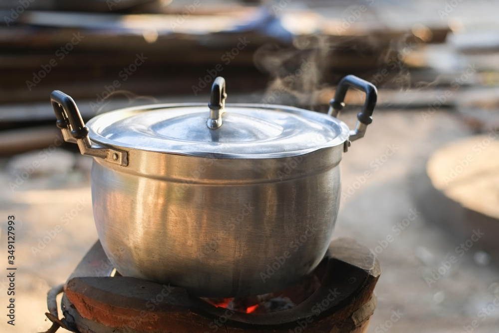 Stainless steel pot that is closed is boiling and has smoke from the steam on the charcoal stove close-up.