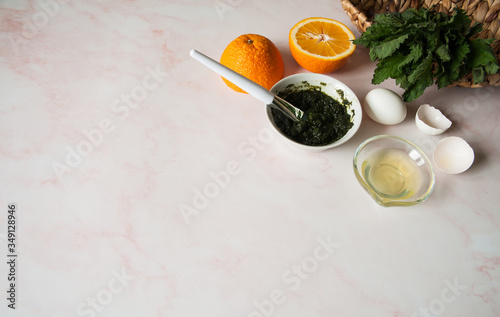 Natural home skin care. Ingredients of a face mask. Crushed nettle leaves, egg white, orange juice and ready-made mask mix in a cup with a spatula on a pastel pink background. Free space.