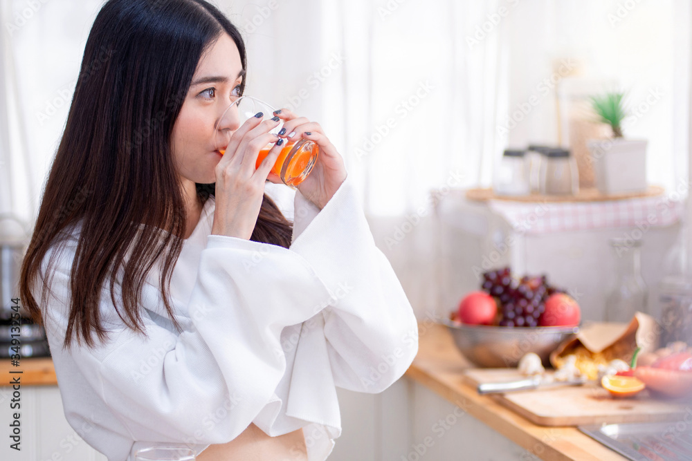 Young Asian woman in the kitchen, Holding with two hands and drinking orange juice in glass, eyes looking forward