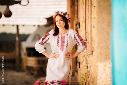 Foto Young redhead moldavian girl dressed in traditional costume with flower wreath on the head standing near old wall