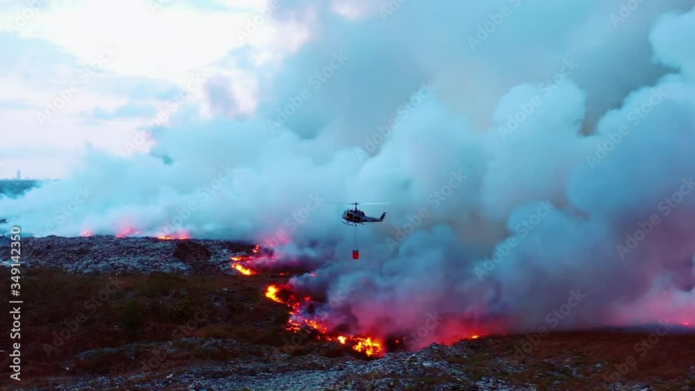Helicopter water drop on California wildfires, Aerial, drone shot following a copter fighting a fire, surrounded by thick smoke, gloomy evening dusk in Los Angeles, USA