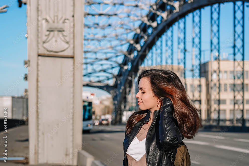 A young business girl in a leather jacket and with a backpack stands on a metal bridge over the Neva river, against the background of a highway in a Sunny beautiful sunset