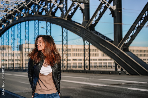 A young business girl in a leather jacket and with a backpack stands on a metal bridge over the Neva river, against the background of a highway in a Sunny beautiful sunset