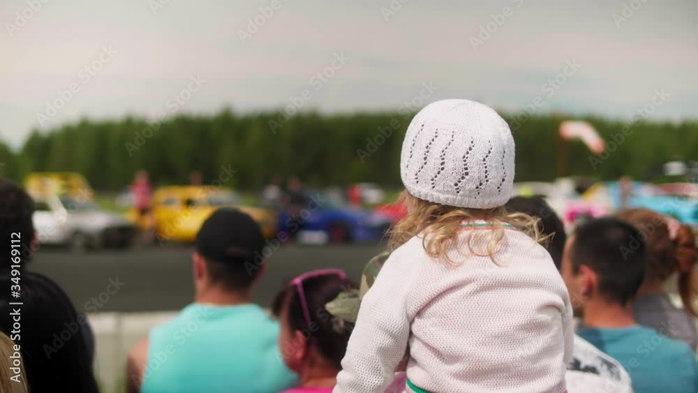 Little girl sits on parent's shoulders in front of crowd and looks at car show Little girl sits on parent's shoulders in front of crowd and looks at car show