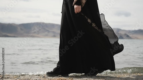 caucasian girl walks along lake shore, in black mourning dress,woman walks on rocks, near mountains