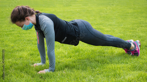 Sport during quarantine, coronavirus, covid-19 concept.Young exercising fitness woman in medical protective mask doing plank or pushups on green grass in park.Caucasian girl training crossfit 