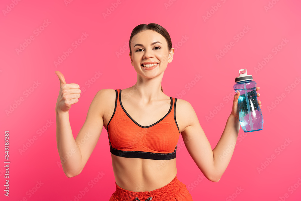 happy sportswoman showing thumb up and holding sports bottle with water isolated on pink happy sportswoman showing thumb up and holding sports bottle with water isolated on pink