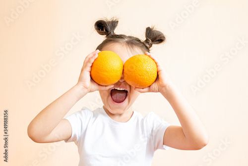A little girl in a white T-shirt holds two orange oranges in her hands. A girl in a white T-shirt laughs and holds two oranges in her hands. A child in a white T-shirt holds two oranges on a beige bac