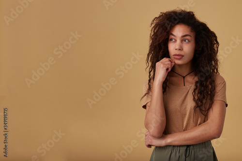 Pensive young pretty curly dark skinned brunette lady with casual hairstyle touching her face with raised hand while looking thoughtfully aside, posing over beige background