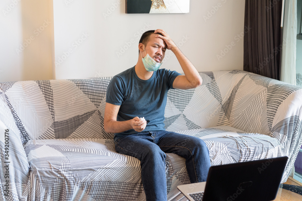 Stressed young Asian man with mask having headache at home under quarantine