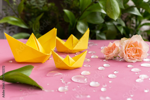 Yellow spring paper ships,boats floating in sparkling water drops on purple background.Pink full roses,branch of green leaves.Summer concept.Greeting card.DIY family activity entertainment with kids