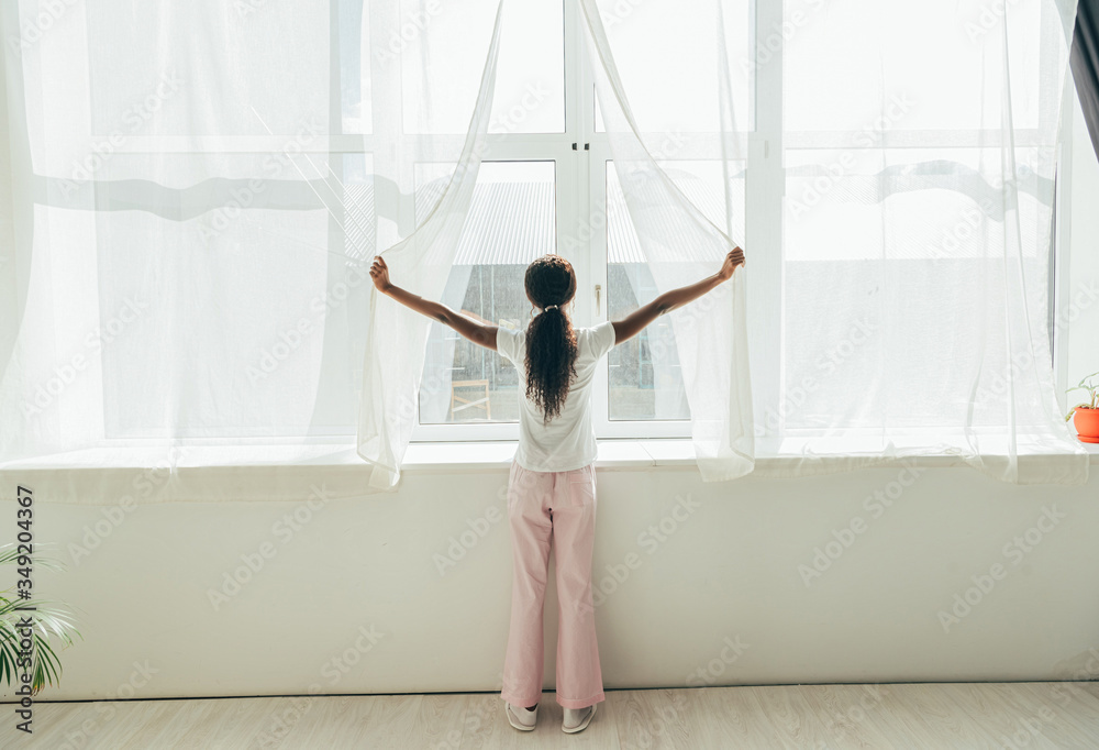 © LIGHTFIELD STUDIOS - back view of african american girl in pajamas opening window curtains in sunlight
