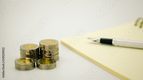 Closeup stack of coins near pen on note book and  white background with copy space. Using as business, financial, money and cooperation concept.
