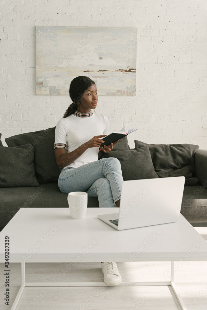 thoughtful african american freelancer holding notebook while sitting on sofa near table with laptop