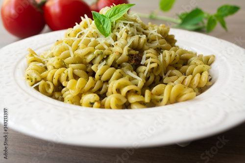 Traditional Italian vegetarian pasta fusilli with fresh homemade basil and pine nut pesto sauce and topped with grated Parmesan cheese. Tomato, basil, parmesan cheese, napkin and fork in background