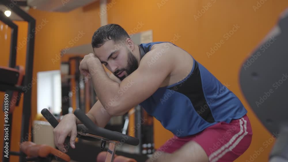 Close-up of exhausted sportsman pedaling exercise bike. Side view of tired young Middle Eastern man riding equipment in gym. Health, lifestyle, sport, endurance, training.