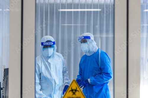 Team of medical doctors in quarantine room check progress with patients. Medical staff wear sterile PPE suit and face shield. Protective hygiene quarantine room with clear COVID-19 sign