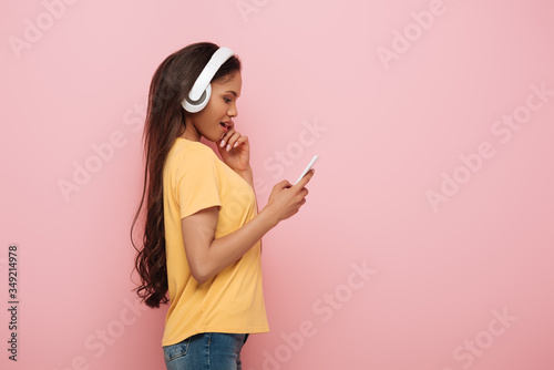 side view of thoughtful african american girl in wireless headphones using smartphone isolated on pink
