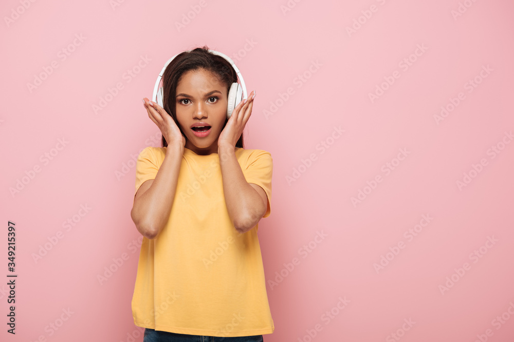 shocked african american girl touching wireless headphones while looking at camera on pink background shocked african american girl touching wireless headphones while looking at camera on pink background