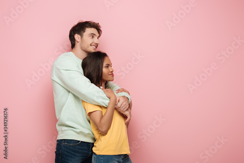 happy guy embracing smiling girlfriend while looking away together on pink background
