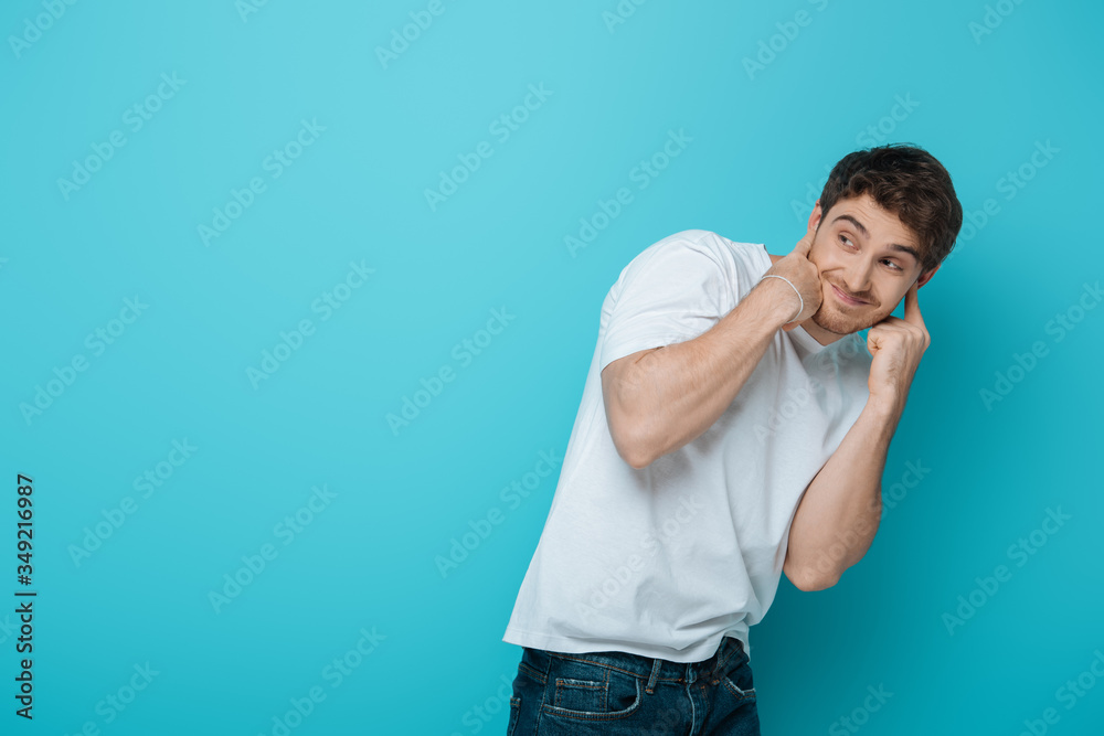 displeased young man plugging ears with fingers while looking away on blue background displeased young man plugging ears with fingers while looking away on blue background