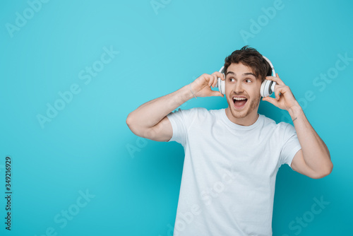 excited guy touching wireless headphones and looking away on blue background