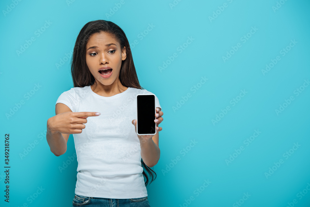 shocked african american girl pointing with finger at smartphone with blank screen isolated on blue shocked african american girl pointing with finger at smartphone with blank screen isolated on blue