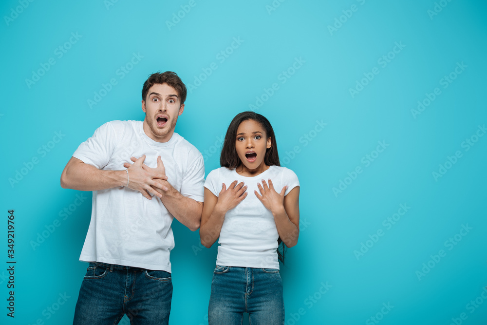 scared interracial couple touching chests while looking at camera on blue background scared interracial couple touching chests while looking at camera on blue background