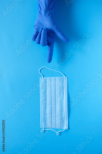 A hand in a protective medical glove points a finger at a protective medical mask. Protect from coronavirus concept on blue background