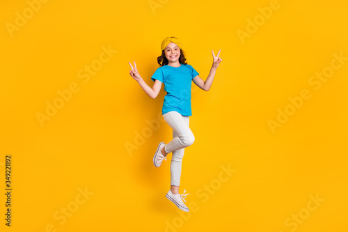 Fototapeta Naklejka Na Ścianę i Meble -  Full body photo of funny little lady jumping high good mood cheerfully meet friends say hello show v-sign wear casual blue t-shirt headband trousers shoes isolated yellow color background