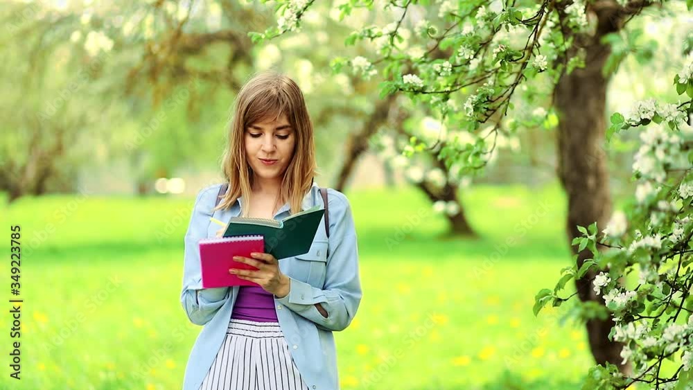 European girl student with long blond hair dressed in a blue shirt, lilac t-shirt writes in a green notebook in the Park. White flowering trees in the background. Concept: education. European girl student with long blond hair dressed in a blue shirt, lilac t-shirt writes in a green notebook in the Park. White flowering trees in the background. Concept: education.