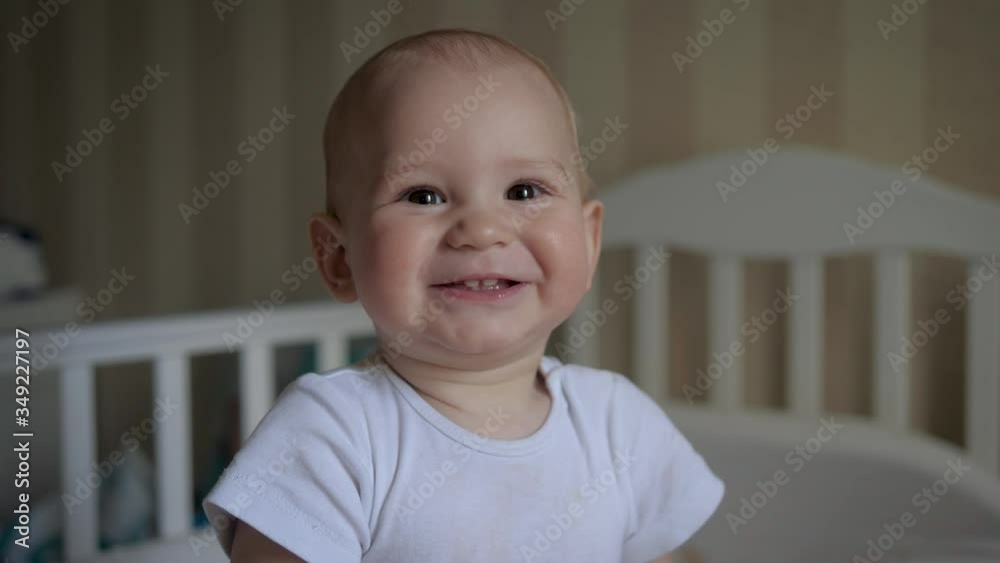 Portrait Of Baby Looking At Camera Smiling Against Background Of Crib In Room Portrait Of Baby Looking At Camera Smiling Against Background Of Crib In Room