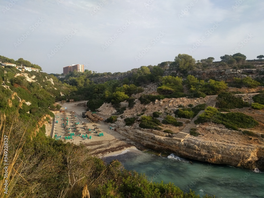 View from the high rocky shore to the sandy beach in the bay of Cala Antena on the island of Mallorca, Spain. A deserted beach with parasols and beach chair in the rays of the rising sun.
