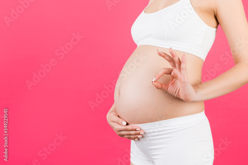 Close up of happy pregnant woman in white underwear showing okay gesture over her abdomen at pink background. Positive pregnancy. Copy space © sosiukin