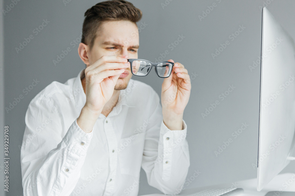 Young man in white shirt looking through glasses. Worker at the desktop with computer on grey background Young man in white shirt looking through glasses. Worker at the desktop with computer on grey background