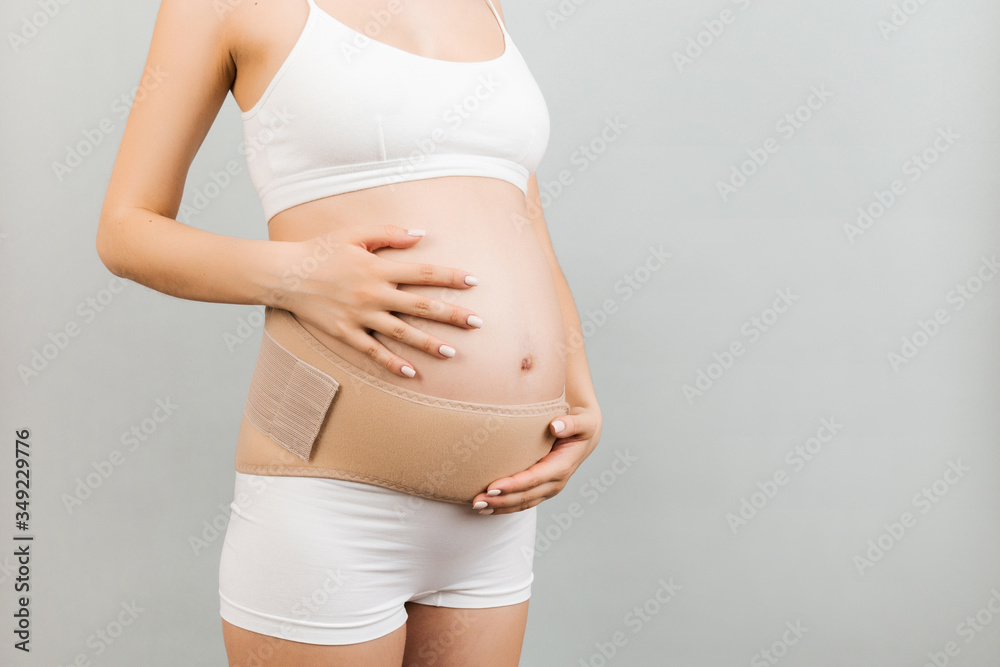 Close up of pregnant woman in underwear using pregnancy belt at gray background with copy space. Orthopedic abdominal support belt concept