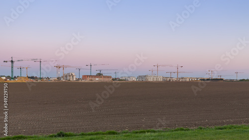 Dźwigi stojące pomiędzy niedokończonymi budynkami w trakcie budowy, za zaoranym polem, na tle wieczornego, kolorowego nieba