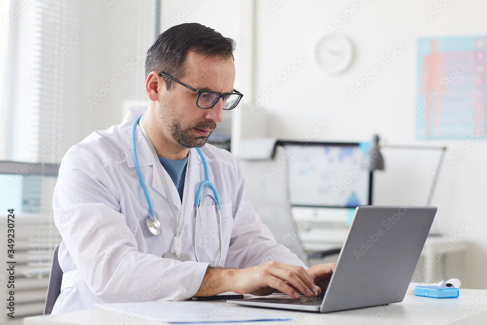 Serious mature doctor in eyeglasses sitting at the table and typing on laptop computer at office Serious mature doctor in eyeglasses sitting at the table and typing on laptop computer at office