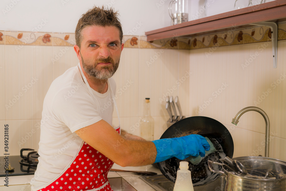 domestic chores stress - young attractive overworked and stressed home cook man in red apron hating dishwashing feeling upset and unhappy washing cooking pan